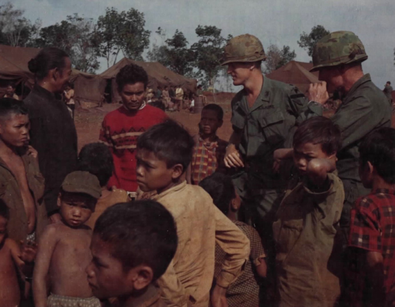 Two American soldiers talking with a highland man surrounded by children in a refugee camp