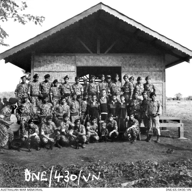 A group of highland soldiers posed in rows in front of a thatched building, with an Australian officer among them