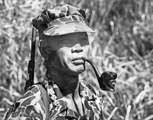 A highland fighter in camouflage and a soft cap, a pipe in his mouth, a rifle slung over his shoulder