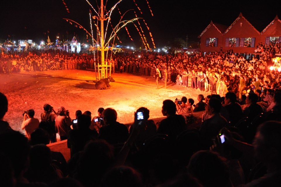 People gathered at night around a fire and a decorated pole at a ceremony in Buon Don