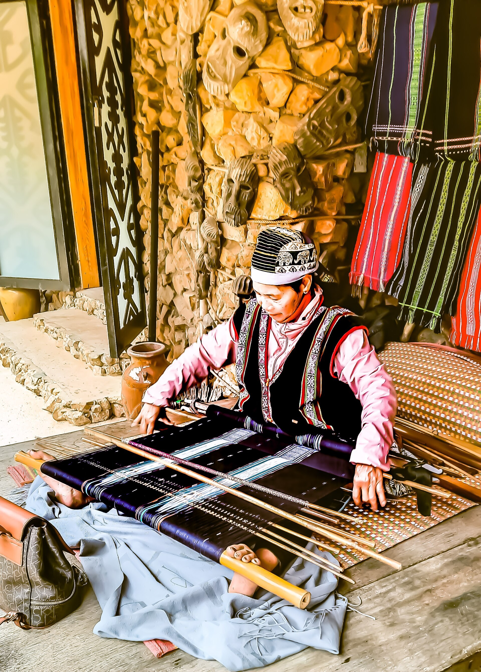 A Koho woman seated on the ground weaving a patterned cloth on a backstrap loom, the warp stretched from her waist to a beam