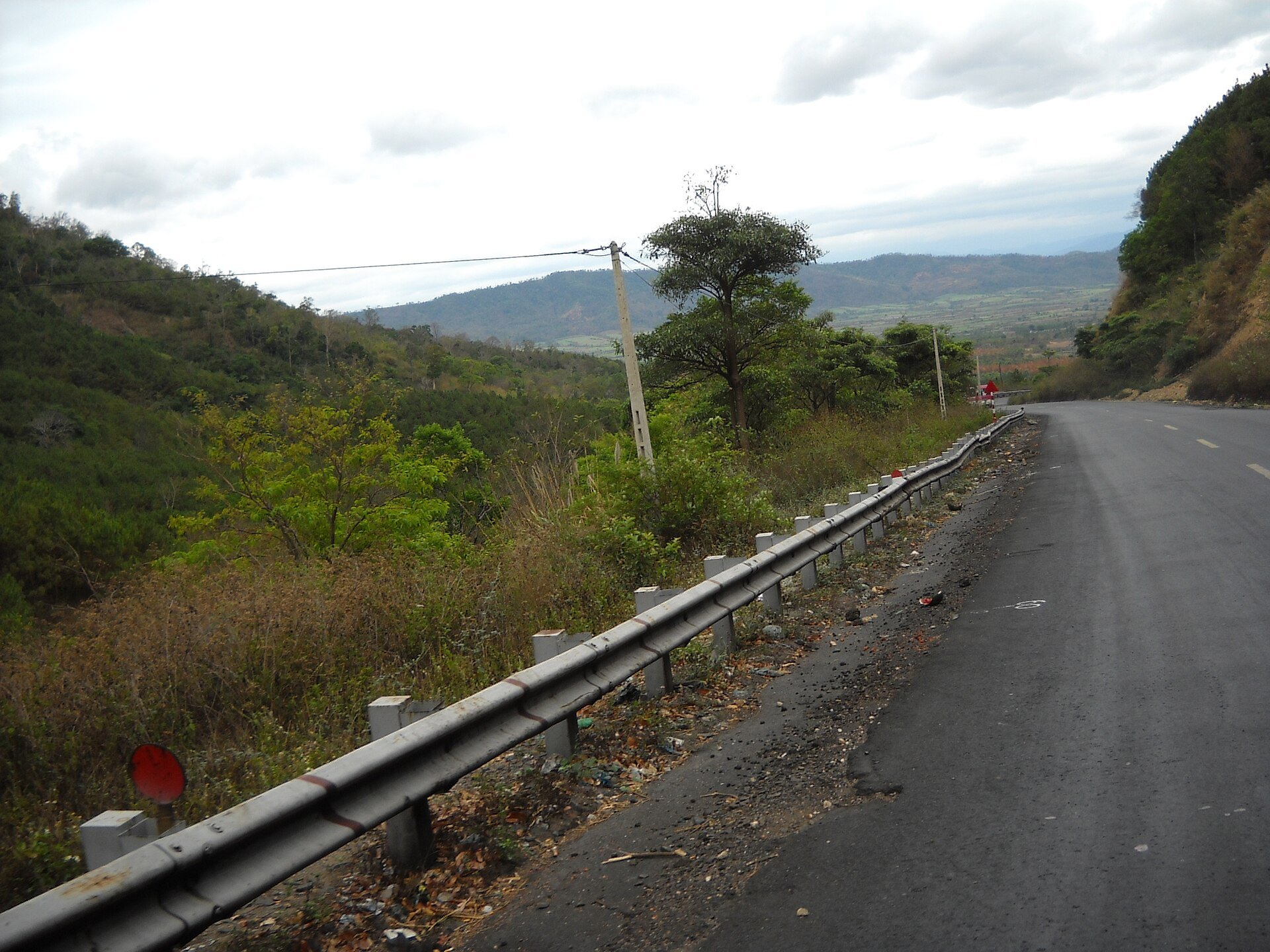 A paved road with a guardrail climbing through dry hills under a cloudy sky