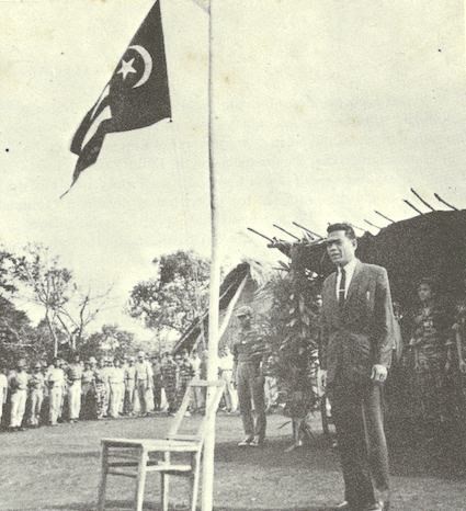 Y Bham Enuol in a suit standing at a flagpole, the flag flying above him, troops drawn up under a thatched shelter behind